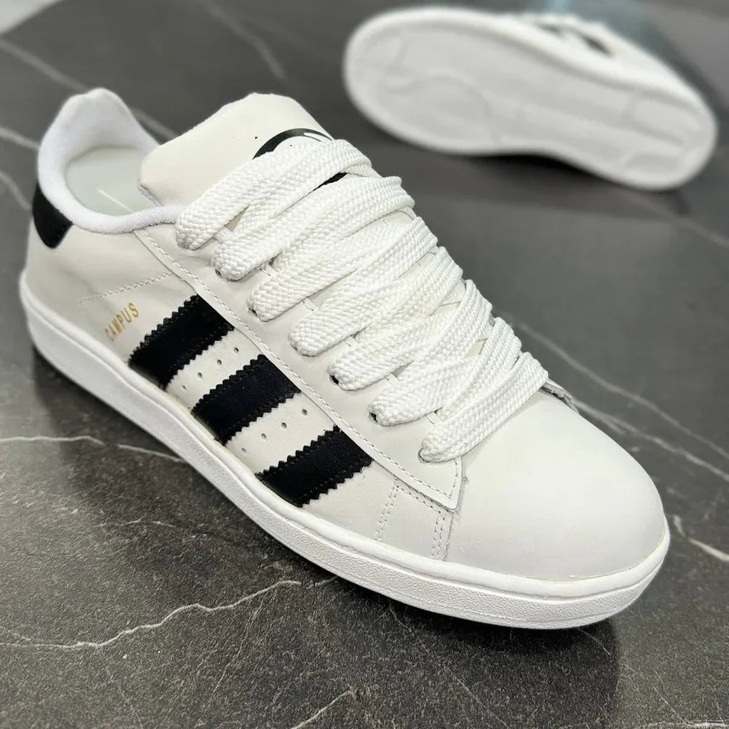 Tênis Adidas Campus branco com listras pretas, cadarço branco, em piso de pedra escura.