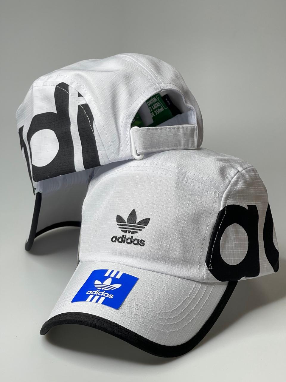 Boné branco Adidas com logo preto, aba curva e detalhe azul sobre superfície cinza.
