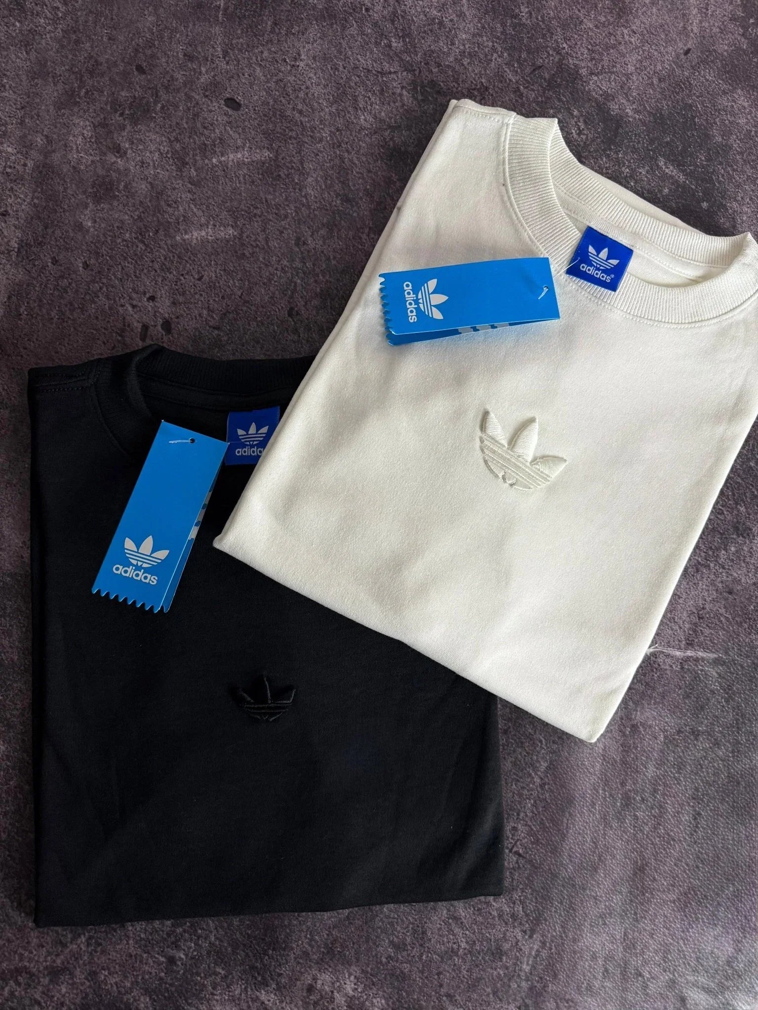 Camisas Adidas oversized preta e branca dobradas, com etiquetas azuis Adidas visíveis