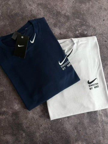 Camisas Nike Swoosh azul marinho e branca dobradas sobre superfície cinza