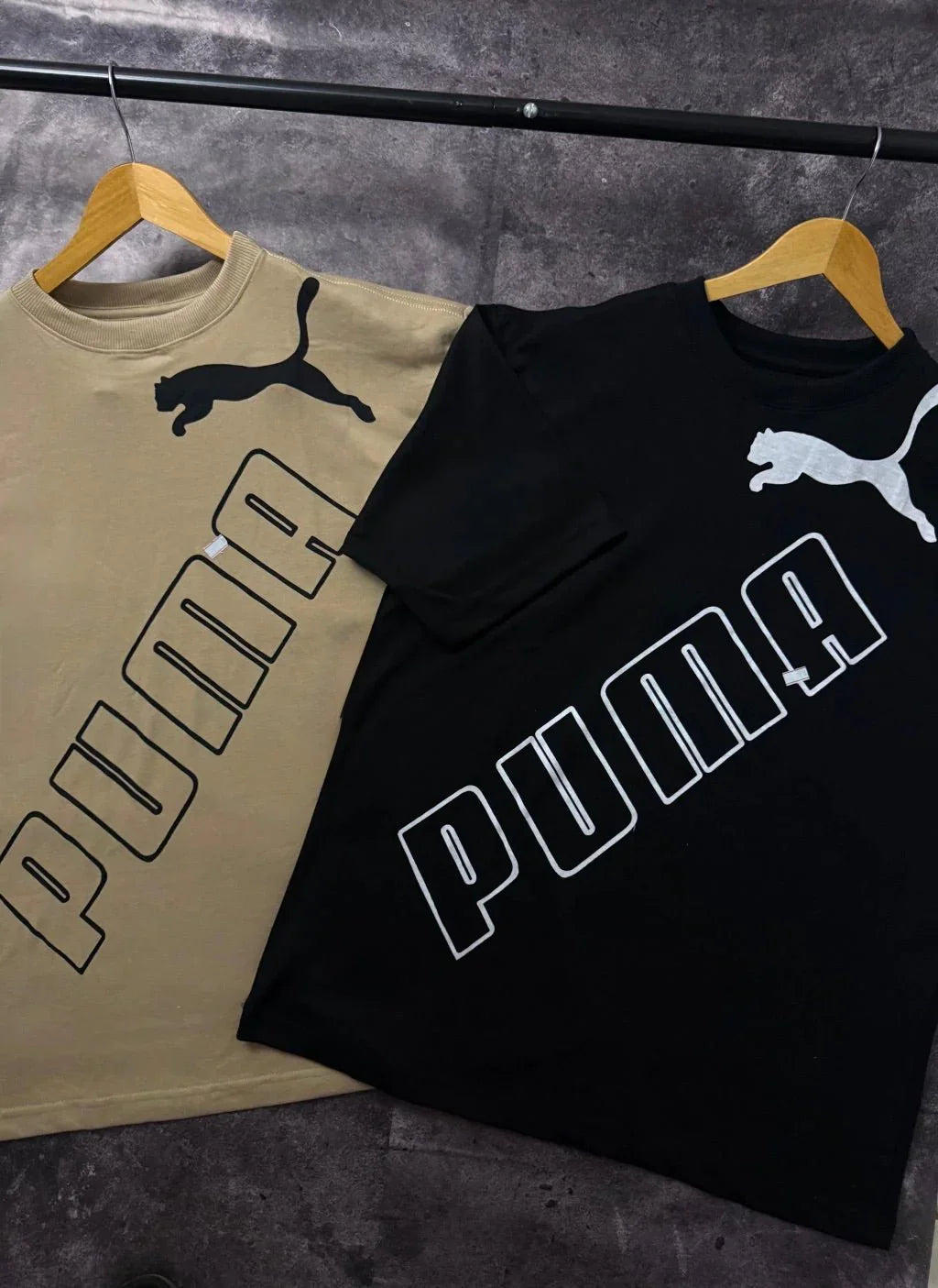 Duas camisetas Puma, uma preta e uma bege, com logo grande e cabides, em fundo cinza.