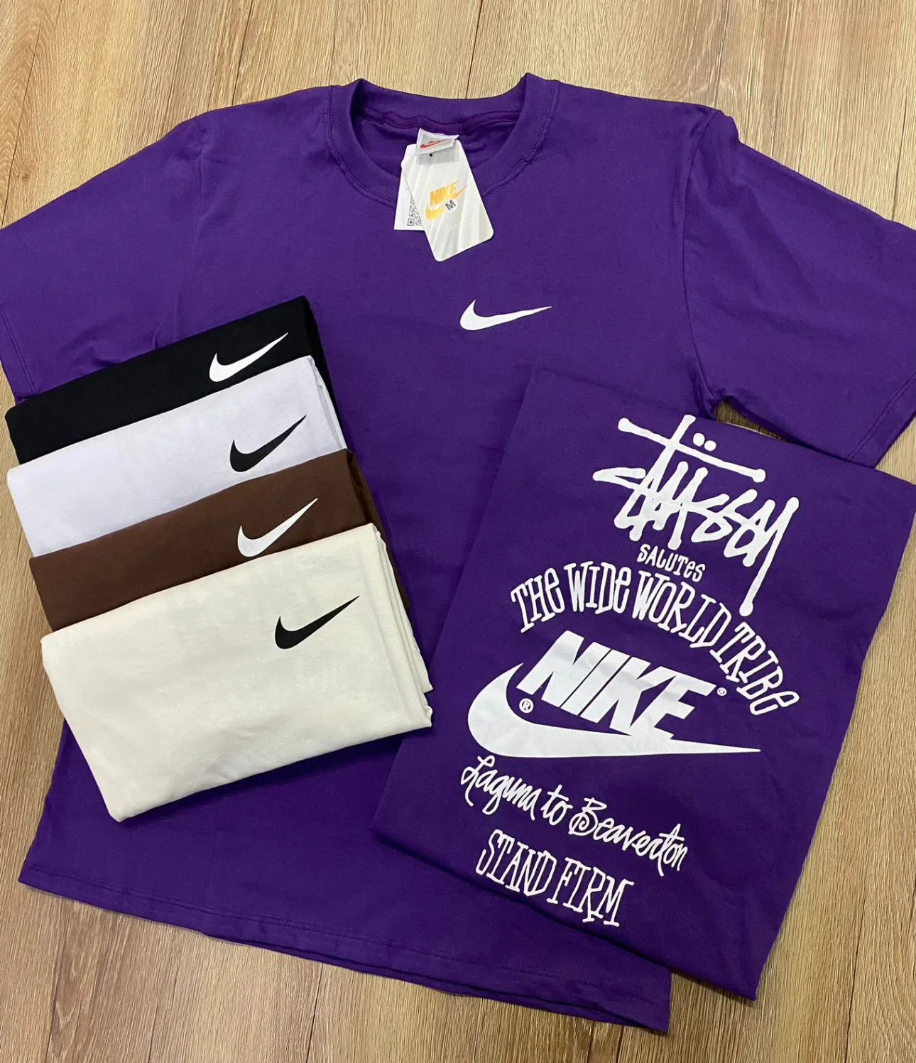 Camisetas Nike e Stussy em várias cores sobre piso de madeira, destaque para modelo roxo.