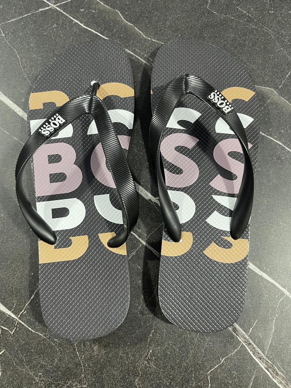 Chinelo Hugo Boss preto com tiras pretas e logo BOSS, sobre piso de mármore escuro.