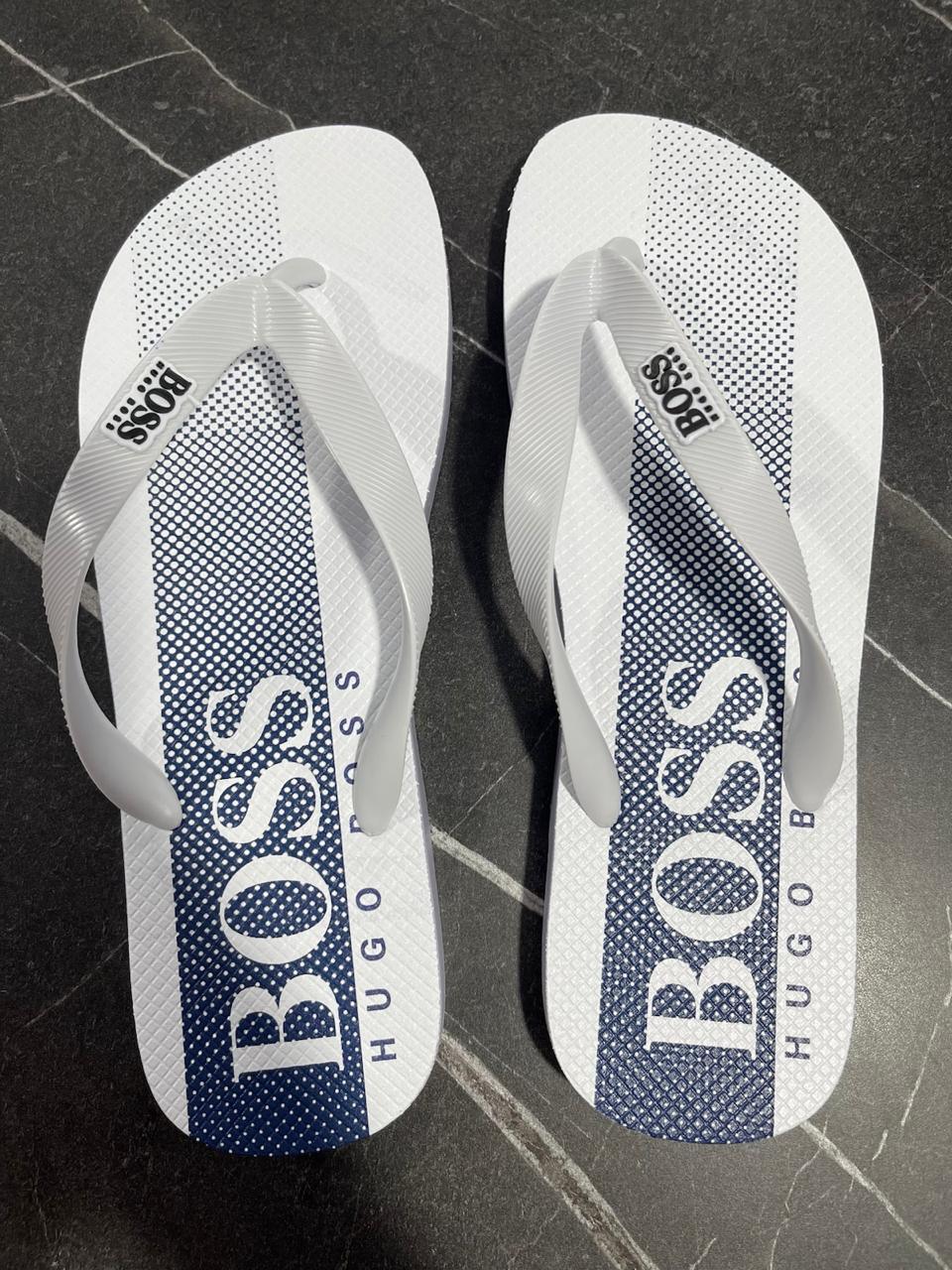 Chinelo Hugo Boss branco com listras azuis e logo BOSS, sobre piso escuro.