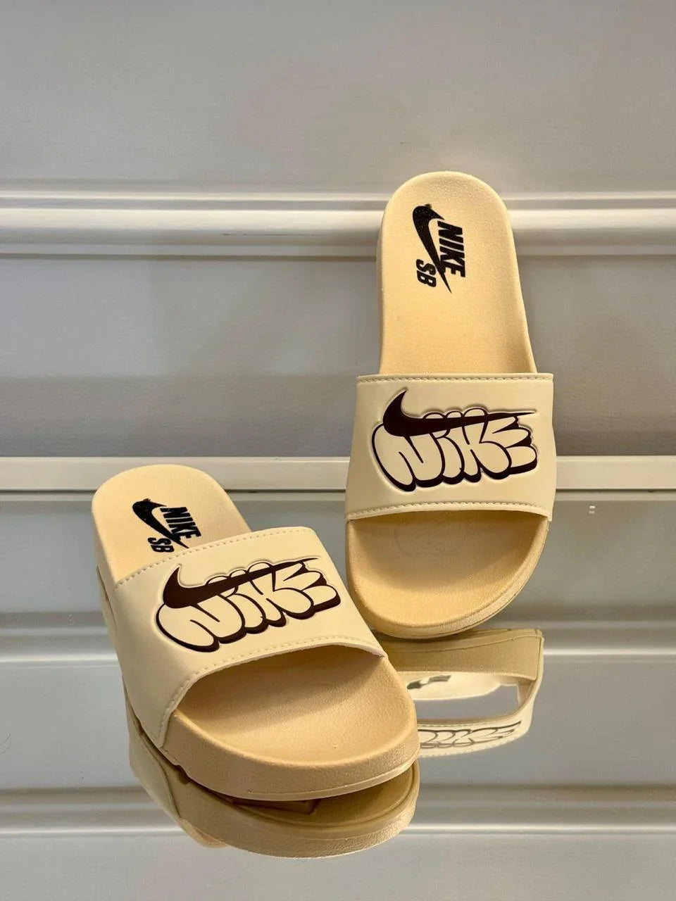 Chinelos slides Nike SB bege com logo preto em destaque, expostos em superfície refletiva.
