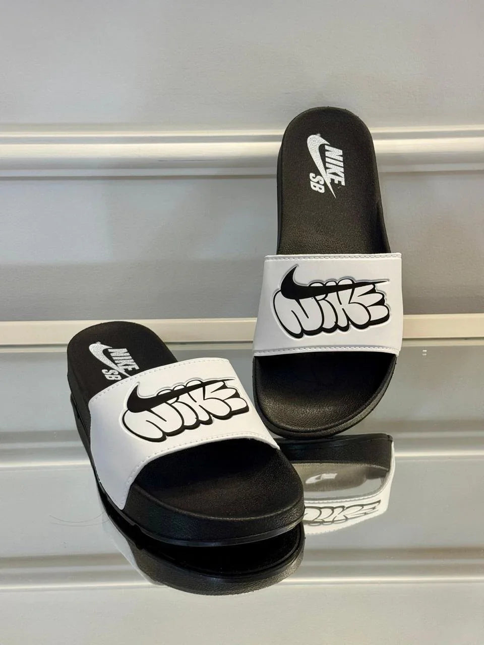 Chinelo slide Nike SB preto e branco com logo grande, exposto sobre superfície refletora