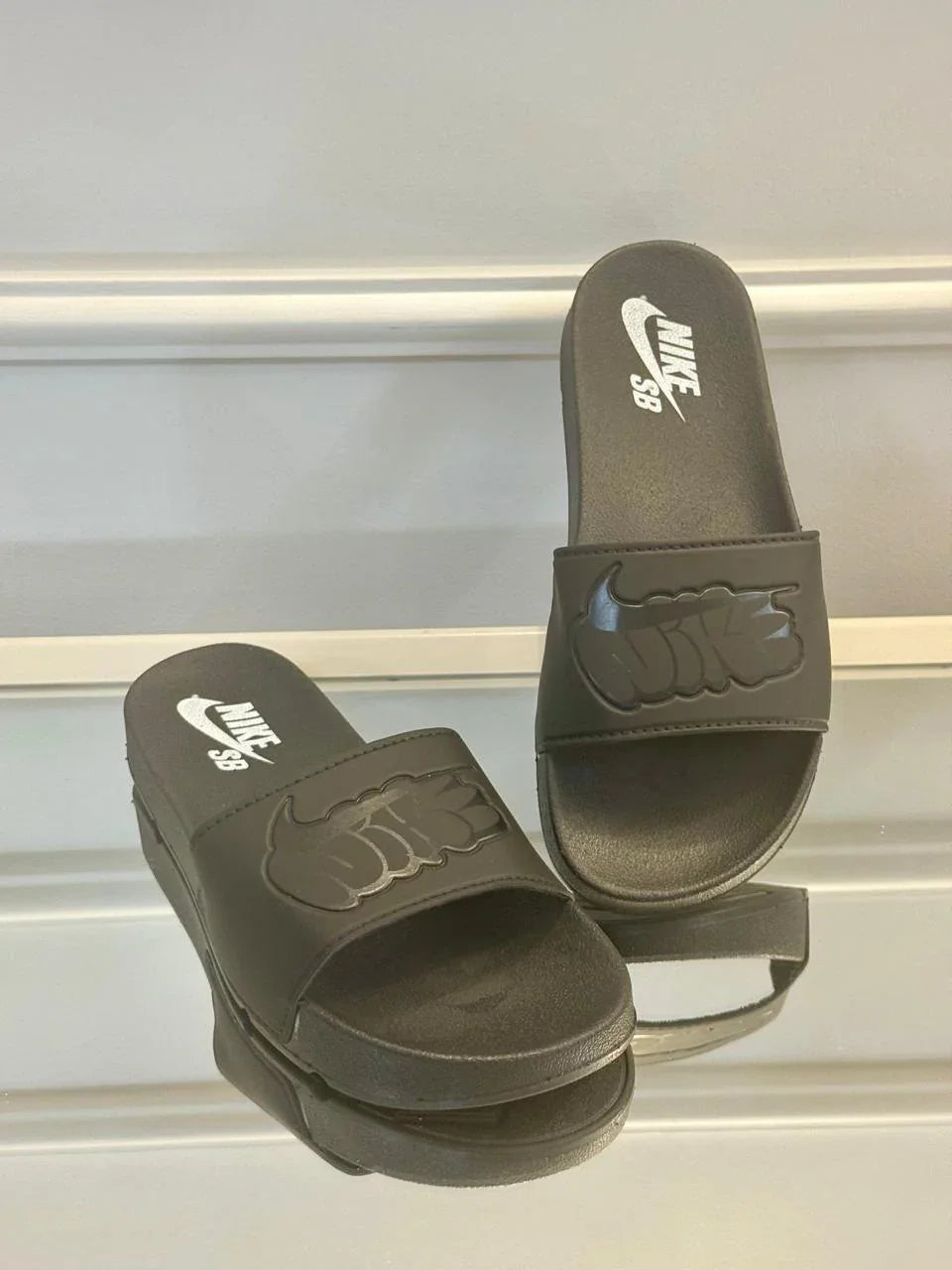Chinelo slide Nike SB preto, logo Nike em relevo, exposto em superfície reflexiva