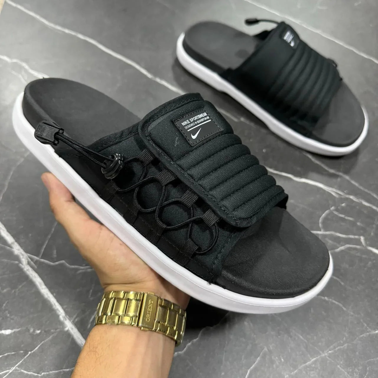 Chinelo Nike Asuna 2 preto com solado branco em mão, exibido em fundo de mármore.