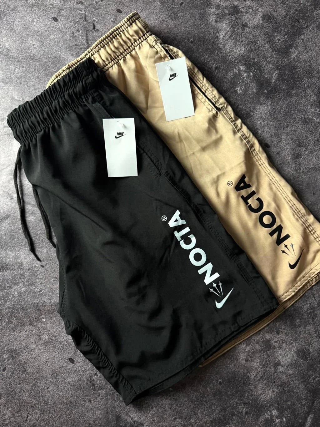 Bermudas NOCTA Nike preta e bege com etiquetas sobre fundo cinza
